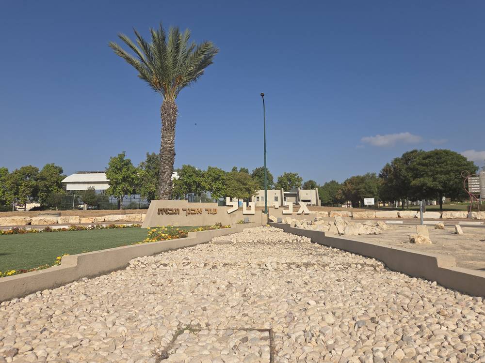אנדרטת גני טל בכיכר הכניסה, ועליה הכתובת עוד אבנך ונבנית, עם דקל ומבני הציבור ברקע